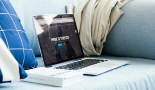 A laptop sitting next to a book on a couch.